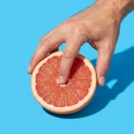 Mano de hombre estimulando el centro de un pomelo rosado cortado, ilustrando cómo estimular el clítoris con los dedos